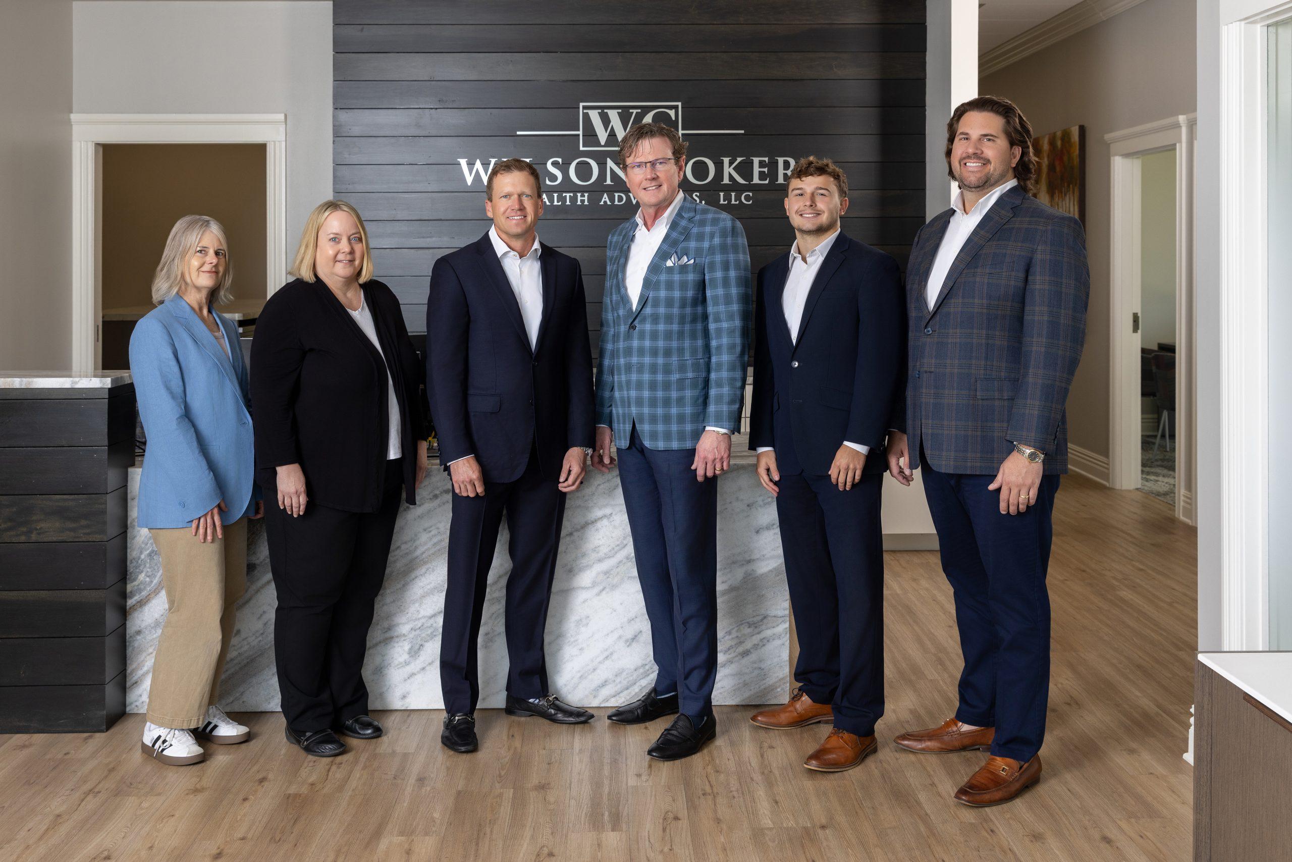 WilsonCoker-083-RET - Wilson Coker Wealth Advisors Six professionals in business attire pose for a group photo in a modern office lobby with a dark wood wall behind them and a sign.